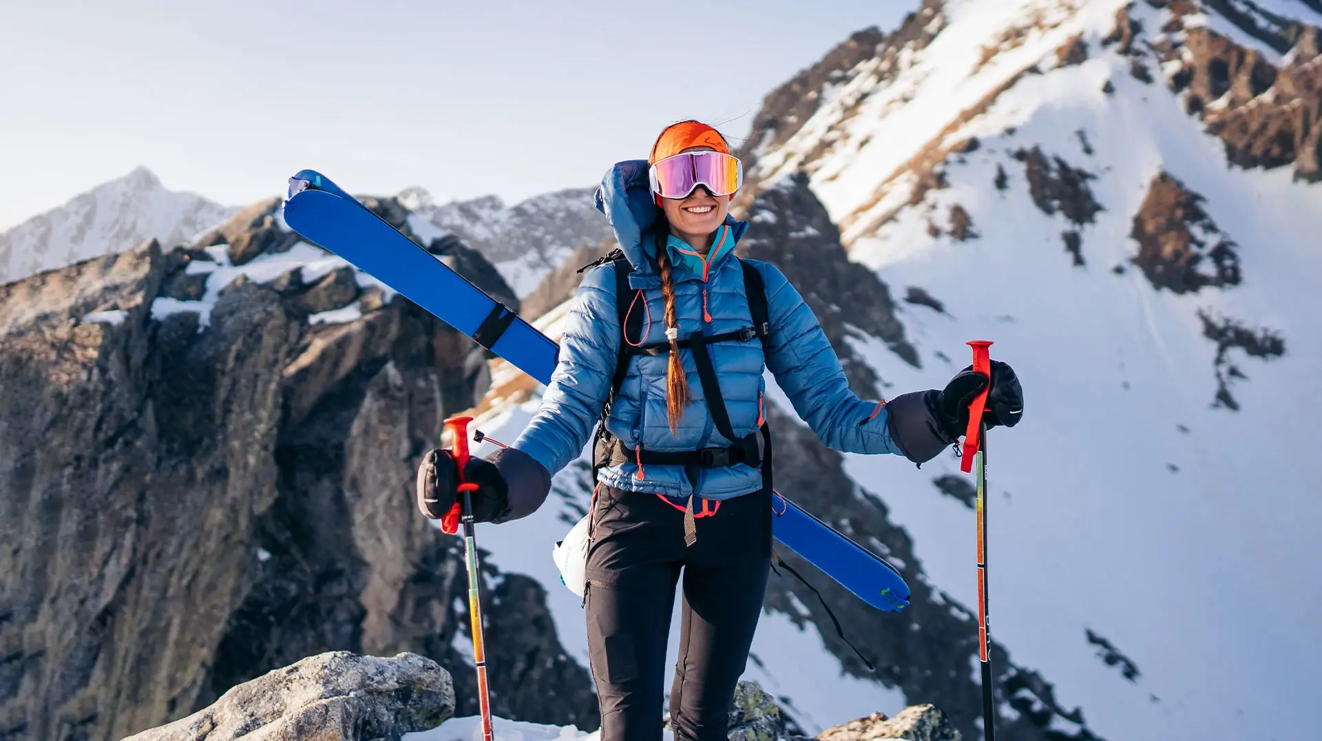 Woman in blue packable jacket with black fitted snowpants wearing orange head wrap big googles, holding ski poles, skis on back of backpack standing on top of a high mountain