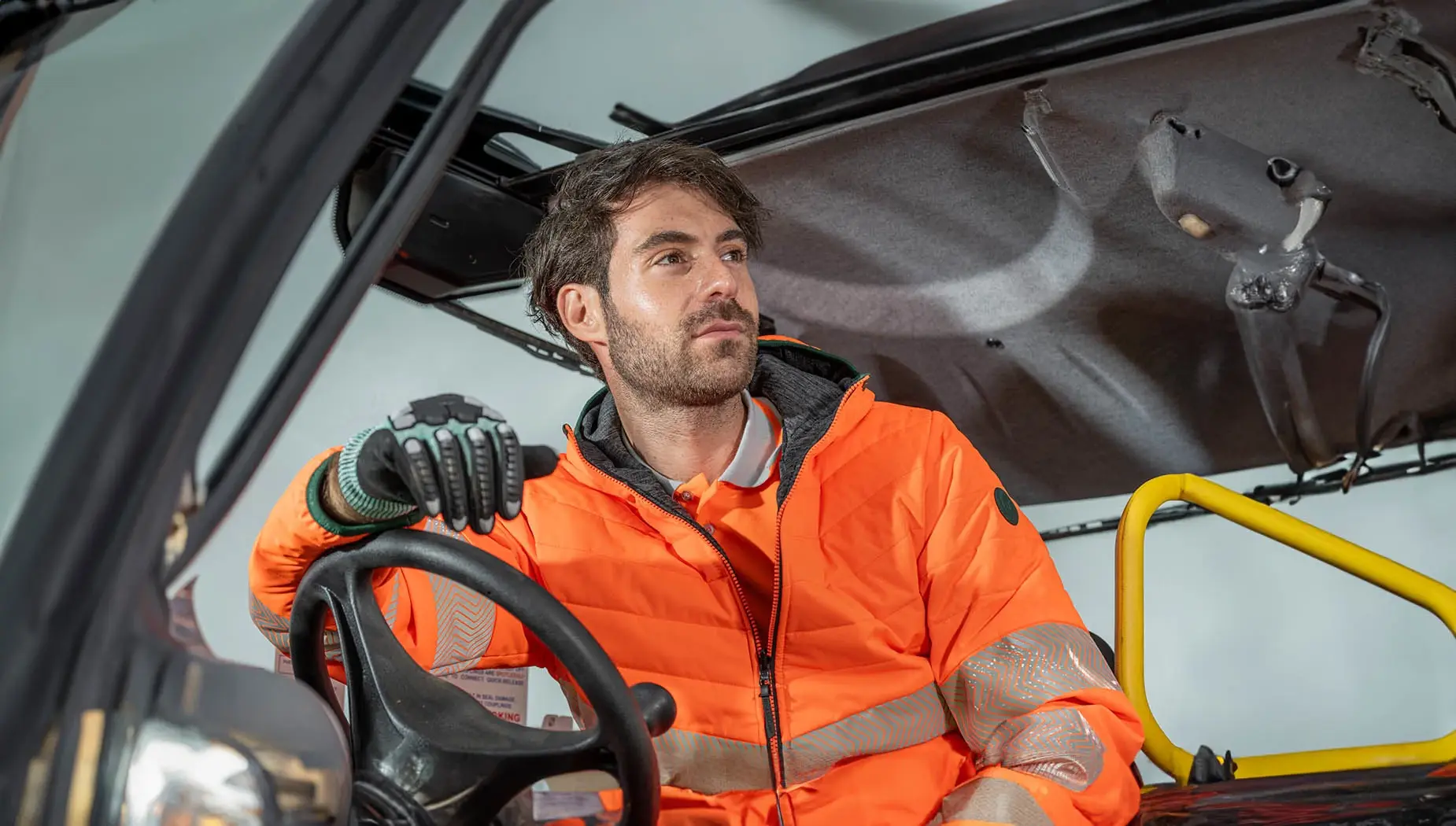 A model sits behind a steering wheel and wears a high-visibility jacket in orange from PULSAR®